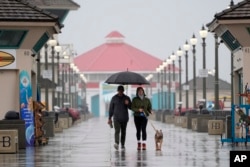 แฟ้มภาพ - คู่รักคู่หนึ่งเดินเล่นพร้อมสุนัขท่ามกลางฝนที่ตกอยู่ที่หาดฮันทิงตัน รัฐแคลิฟอร์เนีย เมื่อ 6 ก.พ. 2567